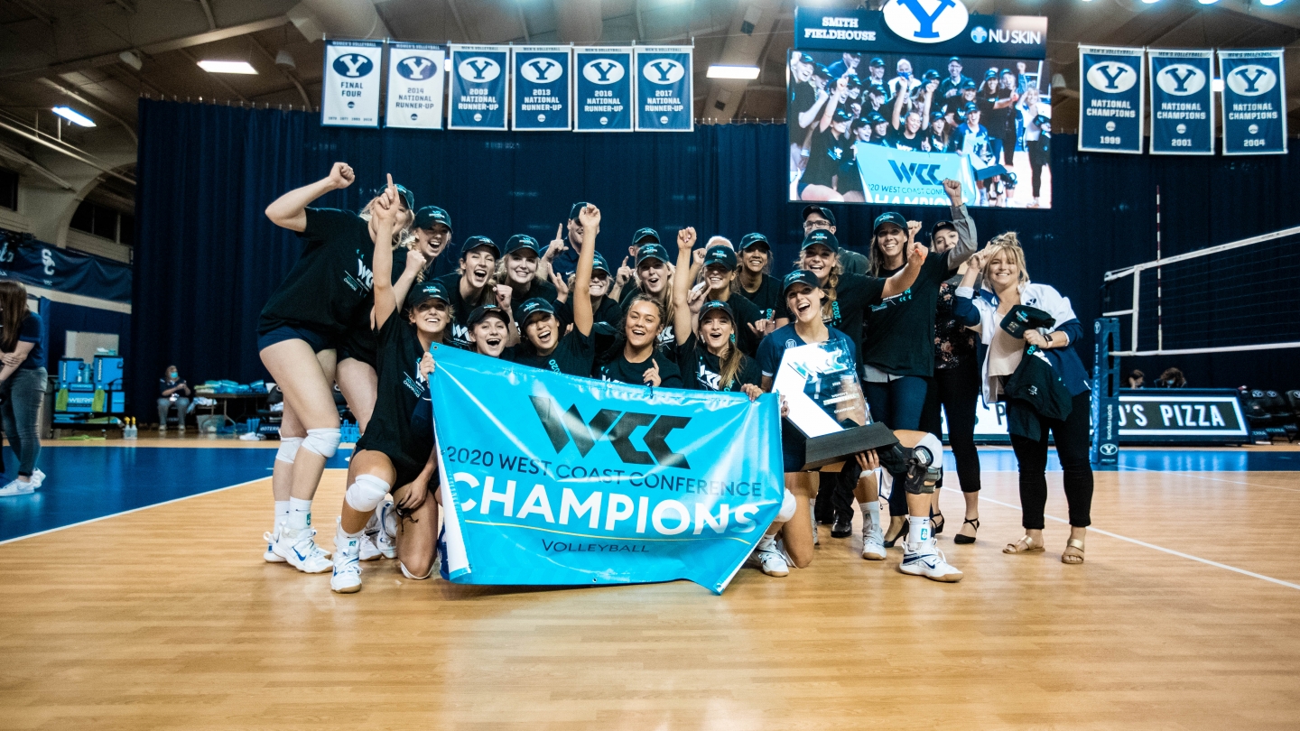 Women's volleyball holds up WCC champion banner and trophy with fists up 