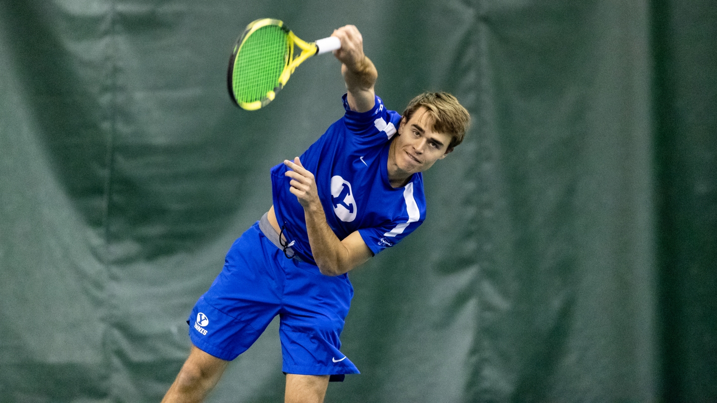 Tennis player in blue uniform serves the ball over his head. 