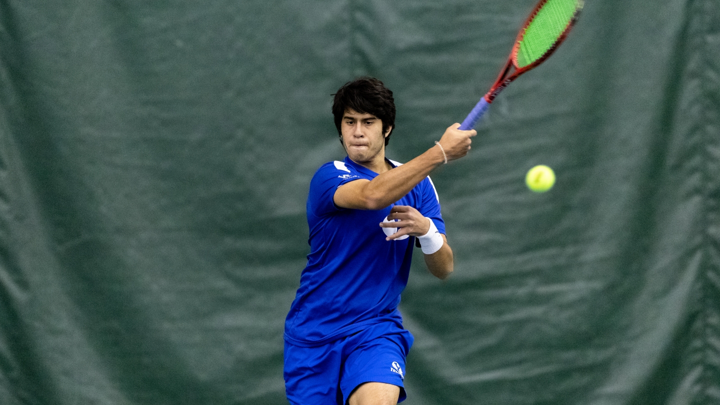 Tennis Player in a blue uniform swings his racquet at the ball using a forearm swing.