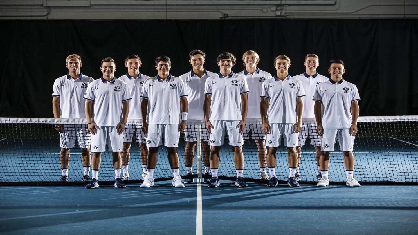 Team photo of the BYU men's tennis team. The players are lined up in front of the tennis net. 